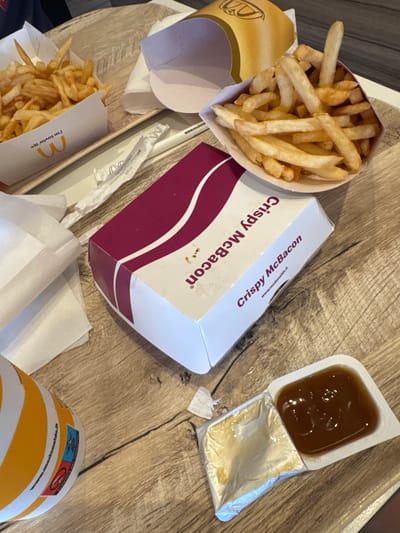 This media file displays a top-down view of a wooden table with several McDonald's food items. The primary subjects are two containers of french fries, one partially visible on the left and another more prominent in the lower left. A box labeled "Crispy McBacon" is positioned in the center, with some text visible including "www.mcdonalds.it". To the right of the box is a small, open container of sauce, likely barbecue sauce given its color. A paper cup with yellow and white stripes, also a McDonald's item, is visible in the lower right corner, with some text and logos on it, including "www.mcdonalds.it" and "PLASTICA NEL PRODOTTO". The overall scene suggests a casual meal, likely lunch or dinner, at a McDonald's restaurant or taken to-go. The lighting appears to be from overhead, suggesting an indoor setting. The image is focused on the food items, with the wooden table surface providing a rustic backdrop. There are no people visible in the image.