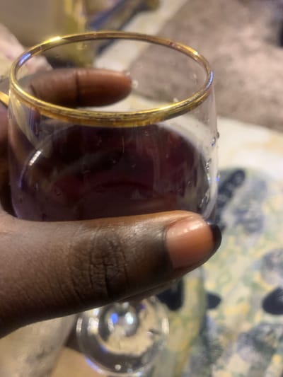 A dark-skinned hand holds a clear glass with a gold-toned rim, containing a deep red liquid, likely a beverage. The exterior of the glass is covered with small water droplets, indicating condensation. The person's thumb is visible, showing a light-colored fingernail with a distinct dark or black tip. The background is significantly blurred, displaying soft gradients of warm and cool colors, with indistinct shapes and textures that suggest an indoor setting with soft furnishings or textile patterns. This scene is situated in Akpajo, Nigeria.