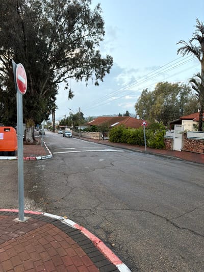 This image depicts a residential street scene in Migdal HaEmeq, Israel, under an overcast sky with patches of lighter blue. The asphalt road surface appears damp and exhibits various cracks and repairs.
In the immediate foreground on the left, a section of reddish-brown brick pavement is bordered by a red and white painted curb. A grey metallic pole rises from this pavement, supporting a circular red traffic sign with a horizontal white bar, indicating "No Entry" (R-202). Partially visible to the left of the pole is a section of an orange public container, possibly a recycling bin. A large tree with dark, dense foliage dominates the upper left quadrant.
The road curves gently to the left and extends into the midground. White painted lines traverse the road surface, consistent with the marking for a speed bump or a pedestrian crossing. Further down the street on the left, two light-colored vehicles, possibly silver or white sedans, are visible either parked or in slow motion.
On the right side of the street, a paved sidewalk runs alongside several single-story residential houses with distinctive red-tiled roofs. These properties are secured by various fencing structures, including stacked stone walls and white metal gates, interspersed with dense green bushes and trees. A red-bordered white triangular road sign, featuring a black symbol resembling a speed bump or road hump, is positioned on a pole on the right side of the road, likely warning of the traffic calming measure ahead.
Overhead, multiple electrical or telephone lines are strung across the scene. The background reveals a landscape of additional residential structures nestled among trees, stretching towards distant hills under the cloudy sky. No individuals or explicit actions or interactions are depicted within the frame.