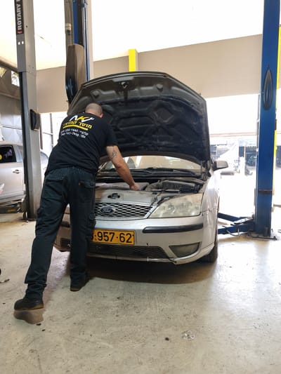 In a garage located in Kafr Yasif, Israel, a man with tattoos on his arm is working on a silver Ford car with its hood open. He is wearing a black t-shirt with a logo and text, and black cargo pants. The car is parked on a concrete floor, and a car lift is visible behind it. The garage appears to be well-lit.