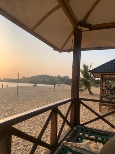 The image presents a serene outdoor scene, viewed from inside a wooden gazebo or shelter overlooking a sandy beach and a body of water. The foreground is dominated by the structural elements of the shelter, including wooden beams and supports that form its roof and side railing. The roof itself appears to be covered with a light-colored, possibly canvas or fabric, material.
Beyond the shelter's frame, a broad expanse of light-colored sand extends towards a calm body of water, likely a river or a bay, given the coastal context of Goderich, Sierra Leone. Across the water, a distant landmass covered with dense green foliage or trees forms the horizon. The sky displays soft, warm gradients of light orange and yellow near the horizon, transitioning to a pale grey or blue higher up, strongly suggesting either sunrise or sunset. The atmosphere appears slightly hazy.
Small figures of people are visible on the sandy area near the water's edge, seemingly strolling or relaxing. A few slender vertical poles are also sparsely dotted across the sand. In the mid-ground, a prominent palm tree stands, and further back, another wooden structure with a dark, possibly tiled, roof is visible, housing more people, hinting at a small beachside facility or restaurant.
The overall impression is one of peaceful leisure and natural beauty. No discernible text is visible in the image.