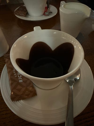 A white teacup sits on a matching saucer, filled with dark coffee that appears heart-shaped. A silver spoon rests on the saucer's edge. A small packet of "Rich Cafe" sugar is placed near the teacup. A similar, empty teacup and saucer are visible to the left of the main set. The scene is set on a wooden surface.