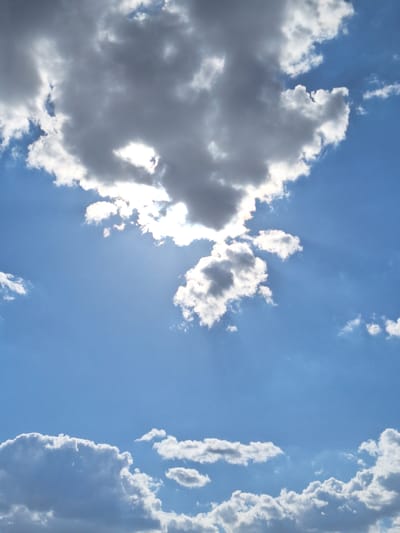 This is a photograph taken from below, looking up at a bright blue sky with scattered clouds. The clouds range from dark gray to bright white, with some appearing to be lit from behind by the sun, which is partially obscured by them. Streaks of sunlight are visible breaking through the clouds, creating a sunburst effect. The clouds are fluffy and varied in shape and size, with some larger formations in the upper portion of the frame and smaller, wispy clouds lower down. The image appears to be taken outdoors in an open area, likely a park or a large, clear space within the city of Markkleeberg, Germany, given the context of the query. There are no people, buildings, or any other objects visible in the frame; the focus is solely on the sky and clouds.