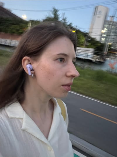 A young woman is seen from the side, looking up and to the right. She has light skin, freckles, and blue eyes. Her brown hair is long and parted to the side, framing her face. She is wearing a white collared shirt with a yellow undershirt visible at the neckline. A small, lavender wireless earbud is in her right ear, and she is also wearing a silver hoop earring.
The background is blurred, but suggests an outdoor setting. There are glimpses of greenery, a building with lights, and a road with a yellow line. The sky appears overcast, suggesting it is either early morning or late evening. The image appears to have been taken in Uijeongbu-si, South Korea. The woman's expression is neutral or slightly pensive. No text is visible in the image.