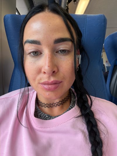 The media file contains a close-up, low-angle, portrait-oriented shot of a young woman lying down. She has dark hair styled in a braid that rests over her shoulder and chest. She is wearing a light pink crewneck sweatshirt over a silver, textured top, and a black choker necklace. Her face is turned towards the camera, with her left eye more prominent. She has a light dusting of freckles across her nose and cheeks. Her lips are full and appear to have a light pink or nude lipstick.
The background is blurred but suggests an indoor setting with blue upholstered seating, possibly on a train or bus. The lighting is bright, indicating daytime. The woman's expression is neutral. There is no visible text in the image. The setting appears to be Ушаки, Russia.