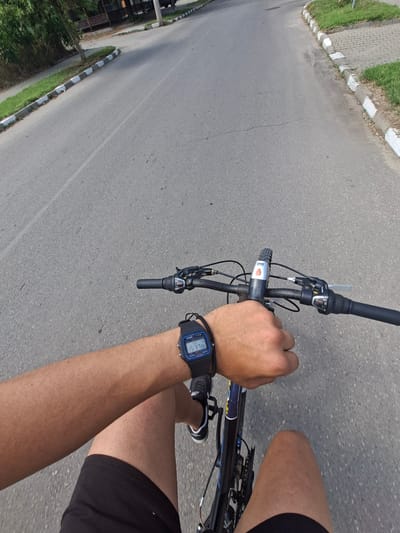 A first-person perspective shot captures a person cycling on a paved road in Galabovo, Bulgaria. The road has a single white lane marking. On the left side of the road, a sidewalk with grass and a white-and-black checkered curb is visible. The right side of the road has a similar sidewalk with paved tiles and grass. The cyclist's left arm is in the foreground, with a blue Casio digital watch displaying "15 37 06" on their wrist. The cyclist's hand is on the handlebar of a black bicycle. The handlebars have a black grip and a silver gear shifter. A silver and red bicycle light is mounted on the handlebars. The cyclist is wearing black shorts. Trees and some structures are visible in the background on the left side of the road. The overall scene depicts a daytime outdoor activity.