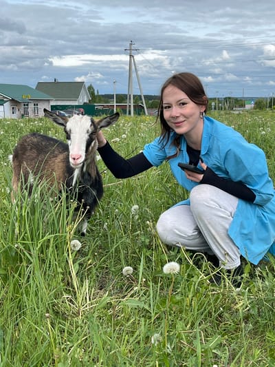The image shows a woman kneeling in a grassy field with a goat. The woman is wearing a blue top, black undershirt, and grey pants, and has brown hair. She is smiling and looking at the camera, her left hand resting on the goat. The goat is brown, black, and white, and is standing in the grass. The background shows a building, power lines, and a cloudy sky. The location is Ludorvay, Russia, identified by the building in the background.
