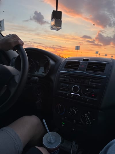 The image captures a first-person perspective from inside a vehicle, looking towards a beautiful sunset or sunrise sky. A person's hand is visible on the bottom part of the steering wheel, indicating they are driving or a front-seat passenger. Part of their leg, wearing gray shorts, is also visible on the right, next to a white and red beverage cup with a straw.
The car's interior features a dashboard and center console. The stereo system has a display showing Cyrillic text: "МУЗ ВТ ВОСПР НАЗВАНИЕ: пл", which translates to "Music BT Playback Name: pl" (likely 'playlist' or 'play'). Various buttons are visible around the stereo, including "MUTE", "RADIO", "AUDIO", "SCAN", and numbered presets (1-6). Below the stereo are climate controls with dials for fan speed, temperature, and air direction. Car keys with a keyring are seen hanging from the ignition on the lower left of the console. An air freshener hangs from the rearview mirror, visible at the top left of the windshield.
Outside the car, the sky is filled with warm, vibrant colors of orange, pink, and purple, indicating either dawn or dusk. Clouds are scattered across the sky. There appear to be reflections or structures of what might be streetlights or poles in the distance. The windshield has some dust or small specks visible, adding to the authenticity of the roadside view in Nagornaya, Russia, as suggested by the provided context and the Cyrillic text on the car stereo. The overall mood is calm and relaxed, perhaps a moment of appreciation for the scenic view during a drive.