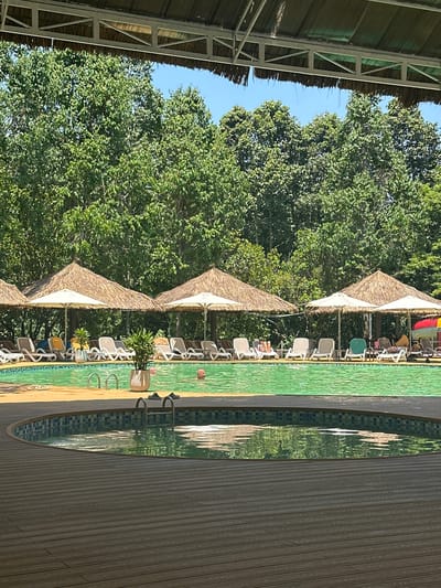 The image shows a resort with a swimming pool and lounge chairs under thatched-roof umbrellas. In the foreground, a wooden deck surrounds a section of the pool. Several lounge chairs are visible along the edge of the main pool, some occupied by people. Two men are in the water, one in the foreground and another further back. Lush green trees form a backdrop behind the pool area, and a clear blue sky is visible above. The scene appears to be a sunny day at a tropical resort.