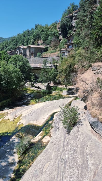 This image depicts a scenic view of the Salt de Sallent waterfall in Rupit i Pruit, Spain. The waterfall cascades over a series of rock formations, with clear, green water flowing into a pool below. Lush vegetation, including trees and bushes, surrounds the waterfall and the rocky terrain. In the background, several stone houses with terracotta roofs are visible, nestled on a hillside. A stone bridge crosses the river, and a paved path with a railing can be seen leading towards the waterfall. The sky is clear and blue, suggesting a sunny day.