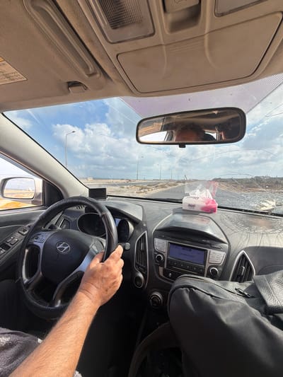The image shows the interior of a car from the perspective of the driver. A person's hands are on the steering wheel, and their arm is visible. The dashboard includes a radio with a display showing "12:45 AUX Connected". A package of tissues is on the dashboard. The windshield is cracked. Outside the car, a road is visible leading to a body of water, likely the sea, under a cloudy sky. The scene appears to be in Nuseirat, Palestinian Territories, based on the context of the request, but this location is not explicitly identifiable from the visual elements alone.