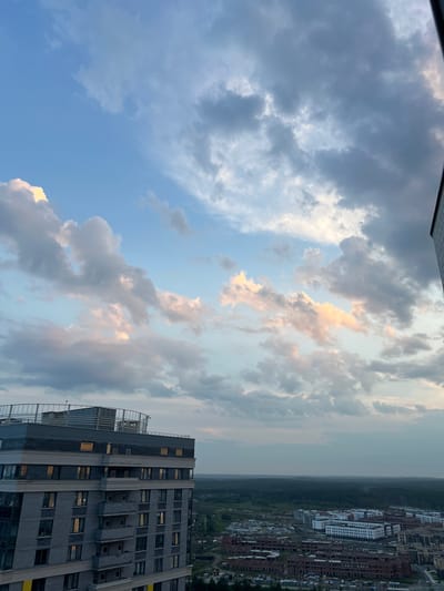 A high-angle view captures a cityscape under a dramatic sky filled with various clouds. In the foreground, a modern, multi-story building with a grey brick facade and numerous windows, some illuminated, dominates the left side of the frame. The building has a flat roof with railings and some ventilation equipment.
Beyond this building, a vast expanse of the city unfolds. Numerous residential buildings, in various stages of construction and completion, are visible. Some buildings are characterized by their white facades and rows of windows, while others are constructed with brick. In the distance, a dense forest stretches across the horizon, meeting the subdued light of the sky. The overall lighting suggests either early morning or late evening. The scene is located in Sovkhozny, Russia. No people or actions are discernible.