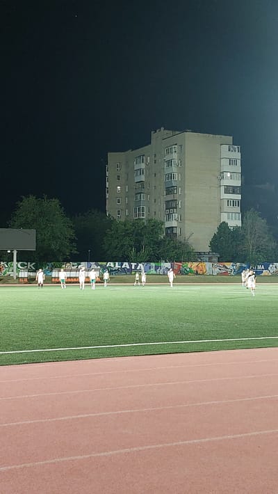 The image depicts a nighttime scene at a football stadium in Novocherkassk, Russia. A group of young athletes, dressed in white and green uniforms, are scattered across the artificial turf field. They appear to be in the midst of a game or training session, with some players walking and others standing still. In the background, a multi-story apartment building looms large against the dark sky, its windows illuminated by interior lights. Trees surround the stadium, and graffiti adorns a low wall in the middle ground. The stadium also features a running track in the foreground, with its reddish-brown surface clearly visible. A bright light source, likely from the stadium's floodlights, illuminates the scene.