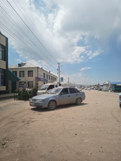 The image captures a sunlit, dusty street scene in Kyzyl-Suu, Kyrgyzstan. A light blue sedan is prominently parked in the foreground, facing left, with its license plate partially visible. Behind the sedan, several other vehicles are present, including a white van and a green truck. Buildings line the right side of the street, some appearing to be businesses with signage in a foreign script. The street itself is unpaved, composed of dirt and small rocks. Utility poles and overhead wires are visible, suggesting a developed, though perhaps not fully modern, urban environment. The sky is blue with scattered white clouds. The overall impression is one of a typical day in a Central Asian town, with daily life unfolding on the street.