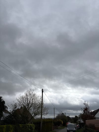 The image depicts a street scene in Longden, United Kingdom, under an overcast sky with dense cloud cover. Power lines run across the upper part of the image, converging towards a utility pole on the right side. Buildings and trees line the right side of the street. Several parked vehicles are visible, including a white van. The road appears to be a narrow residential street.