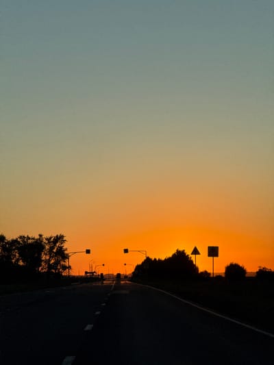 The image shows a desolate road at sunset, leading towards the horizon where the sun is setting. The sky transitions from a pale blue at the top to a vibrant orange near the horizon, suggesting a clear evening. Silhouetted trees line the sides of the road, and several traffic lights are visible, some illuminated, indicating a functioning roadway. Two road signs are prominently displayed on the right side of the road: a triangular warning sign, likely indicating a hazard, and a square sign with a symbol, possibly a speed limit or other regulatory information. The road itself is dark asphalt with white dashed lines marking the lanes. There are no visible people or vehicles in the immediate foreground. The overall impression is one of solitude and an approaching night on a rural or semi-rural road. Without further information, it is impossible to confirm if this scene is located in the city of Морозовка, Russia.
