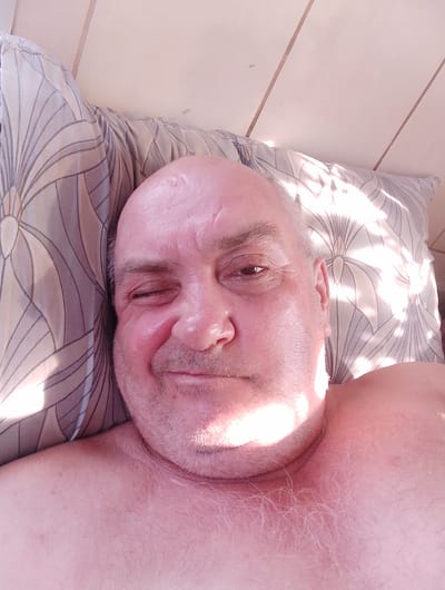 An overhead, close-up selfie shows a light-skinned, bald, middle-aged man lying down, with his head resting on a patterned pillow. The man is looking directly at the camera, with his left eye open and his right eye winking. He is smiling slightly, and his skin appears flushed. The background consists of wooden panels, suggesting an indoor setting. The image is lit by natural light, which creates strong highlights and shadows on the man's face and the pillow. The man is wearing no shirt, and his shoulders and chest are visible, with light-colored chest hair. The image was taken in the city of Krasny Yar, Russia.
