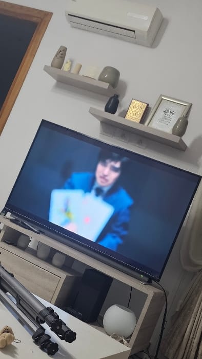 A tilted shot reveals a TV displaying a person wearing a blue outfit holding a white card with red and yellow circular elements. To the TV's left are wall-mounted shelves holding decorative items. An air conditioning unit is also visible on the wall. To the TV's right, there's a light-colored cabinet featuring a rounded lamp, a black speaker, and a tripod. A small, furry object rests on the floor near the tripod.