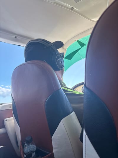 The image depicts the interior of a small aircraft, presumably in transit to or from the vicinity of Chira, Costa Rica. One individual is visible from behind, occupying the pilot's or co-pilot's seat. This person wears a black baseball cap and a black Bose aviation headset. The aircraft's cabin features a light-colored ceiling and side panels. The forward seat exhibits brown, black, and white leather or faux leather upholstery in a geometric pattern. A clear plastic water bottle with a black cap and white label is stored in a pocket on the left side of this seat. A portion of the aircraft's instrument panel, including a circular gauge, is visible to the right of the person's shoulder. Outside the aircraft, through a window on the left, a clear blue sky with sparse white clouds is visible, indicating daylight. A green-tinted section of the windshield or canopy is seen on the upper right. The immediate foreground shows a portion of another seat, upholstered in a similar brown material.