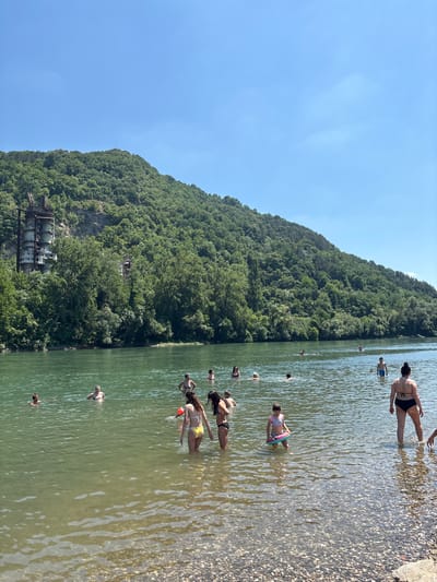 A wide shot captures a clear, sunny day at a river in Zvornik, Bosnia and Herzegovina, with a steep, tree-covered hill dominating the background. The hill features an abandoned, multi-story concrete industrial structure near its base on the left side, with visible metal scaffolding and debris. The river itself is populated with numerous people swimming and wading in the greenish water, some in swimsuits and others with inflatable toys. The riverbed is visible in the foreground due to shallow water and is covered in pebbles. The sky is a bright, cloudless blue.