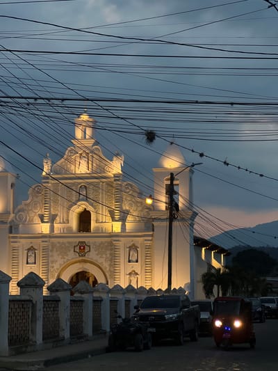 The image captures the Iglesia de San Sebastian in Gracias, Honduras. The white, ornate facade of the church is illuminated, standing out against the dusk sky. Power lines heavily crisscross the view, partially obscuring the church's architecture. To the right, a row of parked vehicles lines the street. A red jeep with bright headlights is visible. The environment suggests a quiet evening in the city.
