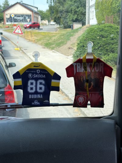 This is a close-up, eye-level shot taken from inside a vehicle, looking through the windshield at Dětmarovice, Czechia. Two decorative air fresheners in the shape of mini sportswear are hanging from the windshield. The air freshener on the left is a dark blue and yellow hockey jersey with the number "86" and the name "BUDINA" printed on it, along with sponsor logos. The air freshener on the right is a red and white shirt-like design with the word "TRAKTOR" across the top and a band of images of people below, and a logo at the bottom. Outside the windshield, a red car is visible on a street next to a building with a sign that is difficult to read. Further down the street, a traffic light sign is visible. Green trees and bushes are present in the background. The interior of the vehicle shows a black dashboard in the foreground.