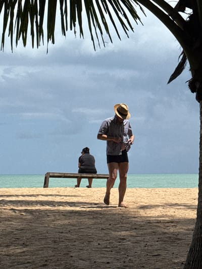 A man wearing a straw hat, checkered shirt, and dark shorts walks on a sandy beach in Ban Khuk Khak, Thailand. He is adjusting the front of his shirt. Further back, a person wearing a dark shirt and shorts sits on a wooden bench, facing away from the viewer, looking out at the ocean. The scene is framed by palm fronds at the top and a palm tree trunk on the right. The sky is overcast and the sea is a calm turquoise. The sand is light brown with scattered dark specks. Shadows from the palm fronds cast stripes across the sand in the foreground.