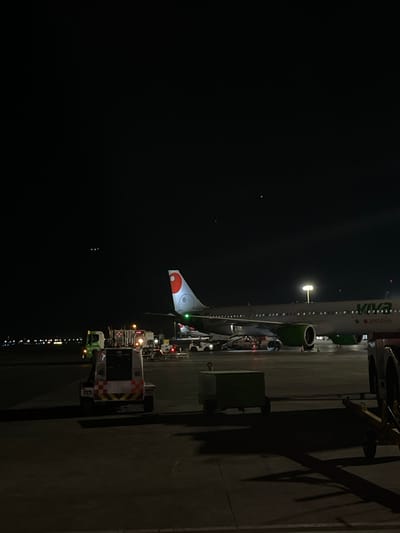 The scene depicts an airport tarmac at night in Apodaca, Mexico. A "Viva aerobus" passenger jet is parked on the asphalt surface, oriented facing right from the viewer's perspective. The aircraft features a gray fuselage with green engine cowlings and "Viva aerobus" branding. Its white tail fin displays a red, orange, and white circular logo.
Multiple pieces of ground support equipment are positioned around the aircraft. In the mid-ground, slightly left of center, a white and orange striped utility vehicle, marked "T10," is present. To its left, a green utility truck with illuminated headlights is visible further back. Other indistinct ground vehicles and cargo loaders are situated near the aircraft's undercarriage and fuselage. A dark green rectangular container rests on the tarmac in the mid-ground, to the right of the striped utility vehicle. Portions of additional cargo dollies or carts are visible in the immediate right foreground.
The area is illuminated by artificial light sources, including a tall pole light illuminating the aircraft and surrounding tarmac, casting shadows. Lights from the ground support equipment and a green navigation light on the aircraft's wing are also visible. Distant airport lights form horizontal lines of illumination on the horizon. The sky above is dark and appears clear. No individuals are discernible within the frame.