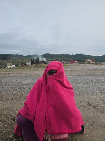 A single individual is observed crouching on a gravel-covered surface in Moqrisset, Morocco. The person is largely enveloped by a bright pink, textured blanket or shawl, which covers the head and body, with dark sunglasses visible over the nose. Portions of a dark, possibly purple, garment and reddish-brown footwear are visible beneath the blanket. The background, beneath an overcast sky, features a scattered urban environment. Various buildings, including light-colored structures and a prominent white building with a red roof, are visible. A distinct plume of white smoke rises centrally in the mid-ground, flanked by two tall, slender trees. Beyond the urban elements, rolling green hills covered with dense vegetation define the horizon.