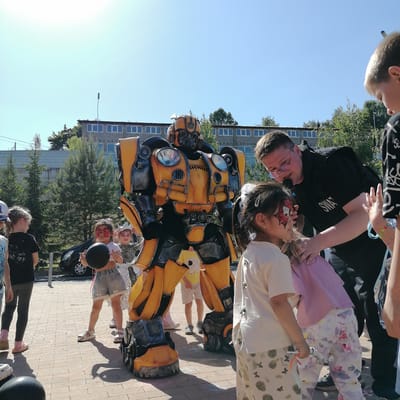 An outdoor scene in Трубачеевка, Russia, features a person dressed as the yellow and black Transformer character Bumblebee standing prominently on a paved area. To the right of the Bumblebee costume, a light-skinned adult male wearing glasses and a black shirt with "SWAT" printed on it is engaging with several children. He is seen closely interacting with a young girl who has red and black butterfly-like face paint, appearing to apply or adjust something on her face. This girl wears a light-colored t-shirt and white and pink floral-patterned pants. Another young girl, also with red and black face paint, is visible to the left of the man, holding a black spherical object and wearing a white crop top and light denim shorts. Further to the left, another child with a silver crown and face paint is partially visible. In the background, a multi-story building with numerous rectangular windows is present under a clear blue sky. Several trees, including some coniferous, are scattered around the paved area. The ground consists of light-colored pavers. Another child, a boy, is partially visible on the far right, looking downwards.