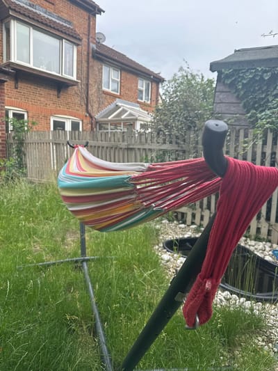 A low-angle medium shot captures a brightly colored striped hammock suspended from a black metal stand in a backyard garden, located in the town of Tewkesbury, United Kingdom. The hammock, featuring stripes of red, yellow, green, white, and aqua, is taut and angled towards the right of the frame. A red rope, likely part of the hammock's support system, is wrapped around the stand's upright pole on the right side.

The foreground is dominated by overgrown, tall green grass, suggesting a lack of recent mowing. Behind the hammock, a weathered wooden fence separates the yard from neighboring properties. A portion of a red brick house with white-trimmed windows is visible to the left, with a bay window on the upper floor. To the right, a small wooden shed with a dark, sloped roof is partially covered in ivy. In the background, a black pond liner or water feature is partially visible amongst white gravel and more greenery. The sky is overcast, indicating a cloudy day. No people are present in the image. The scene depicts a residential garden environment.