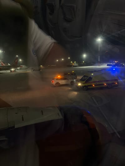 The scene depicts a nighttime airport tarmac at Stolbishche, Russia, viewed from inside an aircraft. The lower right portion of the frame shows a section of an aircraft engine nacelle, indicating the viewer's position. Reflections on the upper left of the window reveal a person's arm in a white shirt. The tarmac is covered in a layer of snow or ice.

In the midground, a white SUV, marked with "229" on its side, is stationary with an amber rotating beacon active on its roof. Adjacent to it, a white ambulance with yellow and red stripes, bearing the number "53" and Cyrillic text, has its amber headlights illuminated. A blue flashing light is visible further behind the ambulance. A white utility van is partially discernible behind the SUV.

In the background, multiple aircraft are parked, including one displaying "REDWING" on its fuselage and tail. Various airport ground service vehicles are present across the snow-covered tarmac. The entire area is brightly illuminated by numerous tall light poles, and additional blue flashing lights are visible in the distance, suggesting further activity or emergency vehicles. No direct interactions between individuals are visible.