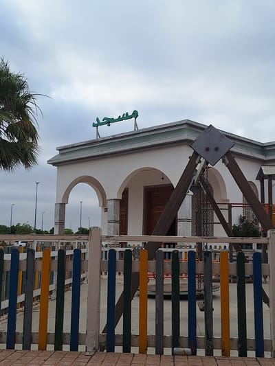 The image shows a view from behind a brightly colored wooden fence in Oulad Hcine, Morocco. The fence has vertical posts painted in alternating colors of blue, yellow, green, and white. Beyond the fence, a white building with arched entrances is visible. Green Arabic script, which translates to "Mosque," is mounted on the roofline of the building. To the left of the building, a portion of a palm tree is visible. A wooden swing set structure stands in front of the building, and to the right, part of a playground slide can be seen. The sky is overcast. The scene depicts a public space that appears to be a mosque with an adjacent children's play area.