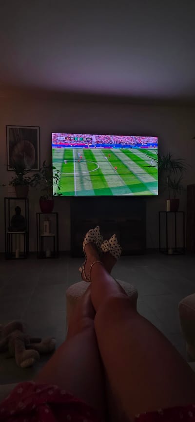 In Lieli, Switzerland, a person is relaxing in a dimly lit living room, watching a soccer match on a large television screen. The room has a modern aesthetic with clean lines and dark flooring. The television displays a live soccer game with a scoreboard indicating the time is 65:54, and the score is 0-0. The person's legs, adorned with white polka-dot high heels featuring large bows, are crossed and positioned in the foreground, suggesting they are seated on a couch or ottoman. A stuffed animal lies on the floor near their feet. The background features a framed picture on the wall, several potted plants on stands, and minimalist decorative items, including a statue. The overall atmosphere is one of relaxed leisure.
