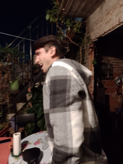 A blurry, poorly lit image depicts a man in a plaid hooded jacket outdoors at night in Gobernador Julio A. Costa, Argentina. The man is facing left, mouth open as if speaking or laughing. Behind him is a patio with plants, a black metal railing, and a brick structure. A table with items, including a roll of paper towels and a black object, is in the foreground.
