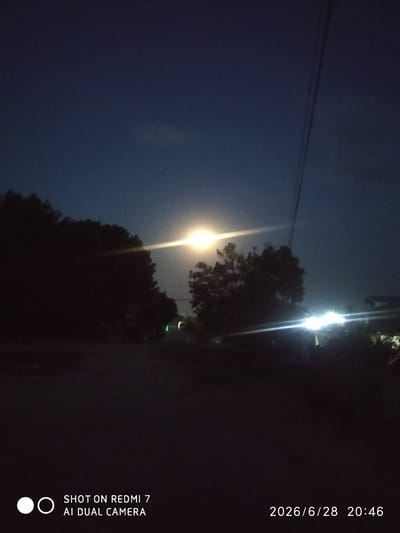The image depicts a nighttime street scene in Khalkabad, Uzbekistan. The sky is a deep blue-gray, suggesting twilight or a moonlit night. A bright light source, possibly a street lamp or the moon, is visible in the upper center of the image, emitting a strong glare. To the left, silhouetted trees form a dark mass against the sky. In the mid-ground and right side of the image, there are other bright lights, indicating buildings or street lighting. A thin power line runs diagonally across the upper portion of the frame. The foreground is dark and indistinct, appearing to be a road or pathway. Text at the bottom of the image reads "SHOT ON REDMI 7 AI DUAL CAMERA" and a date/time stamp "2026/6/28 20:46". There are no people or visible signs of activity in the image.
