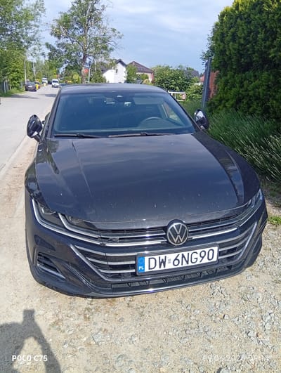 A black Volkswagen Arteon is parked on the side of a gravel road in Wilczyce, Poland. The car is facing the camera, and its front end is visible. The license plate reads "DW 6NG90". The background shows a street with other cars, houses, and trees. The sky is cloudy. The photo was taken on June 9, 2026, at 09:14.