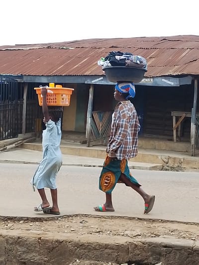 In Iseyin, Nigeria, two individuals walk along a paved street in front of a building with a corrugated iron roof. The person on the left, a child, is wearing a pale blue robe and carrying an orange plastic basket filled with yellow containers on their head. The person on the right, an adult, is wearing a plaid shirt over a white top and a colorful patterned skirt. They are carrying a black basin filled with folded clothing on their head, balanced by a blue and red headscarf. The street is lined with a low concrete barrier on the right side of the image. The building appears to be a storefront, with a sign reading "MIGNER" visible on its facade.