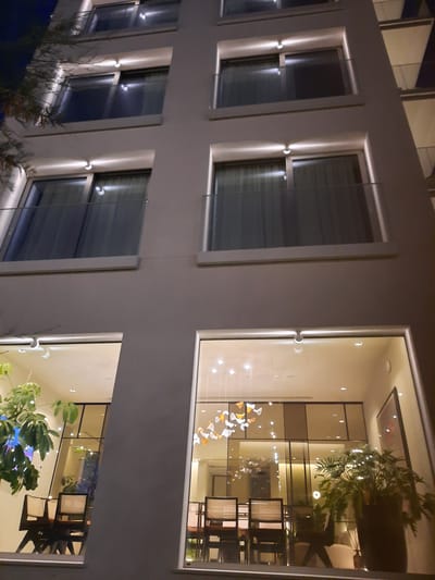 The image shows the exterior of a modern hotel building at night, with lights illuminating the windows and balconies. The building appears to be constructed of light-colored material, possibly concrete or stucco. Several floors are visible, each featuring large windows with glass railings on the balconies. Inside some of the rooms, furniture such as tables and chairs can be seen, along with decorative elements like plants and a chandelier. The overall impression is one of contemporary design and nighttime ambiance. The provided metadata suggests the image was taken in Pesaro, Italy.