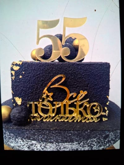 The image shows a close-up view of a dark blue cake decorated with gold accents. On the left side of the cake, large gold numbers "35" are prominently displayed. A gold cake topper with intricate lettering and a heart shape is placed on the front of the cake. The cake itself appears to have a textured surface and is adorned with small pieces of gold leaf. The background is a light color, possibly a table or a surface. The overall presentation suggests a celebration, likely a birthday for someone turning 35. The lighting is bright, illuminating the gold decorations and the textured surface of the cake. There is no visible information about the time of day, weather, or specific emotions conveyed.