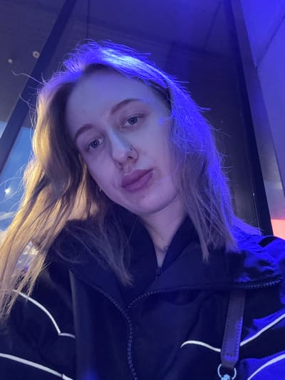 A young woman with long, light brown hair is photographed indoors, likely in Ol'ginskaya, Russia, given the prompt. Her hair is lit with a blue cast at the top of her head, fading into a more natural brown color towards the ends. She is wearing a dark jacket with a zipper and white stripes. Her face is visible, showing light skin, blue eyes, and a piercing in her left nostril. The background is dark, possibly a wall or window at night. The photo is taken from a slightly elevated angle.