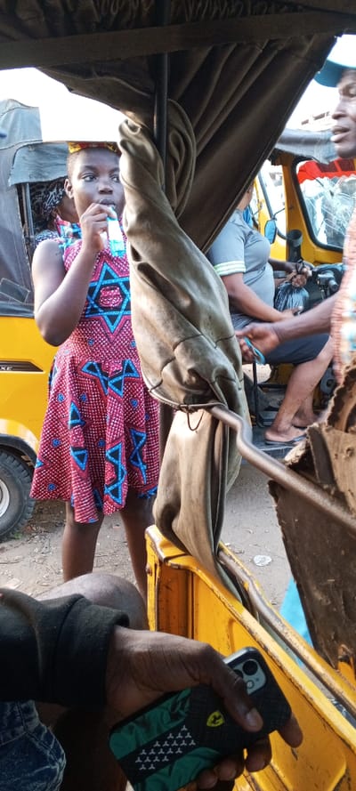 A young person in a red and blue dress drinks from a clear plastic bag. She is outside a yellow vehicle, likely a tuk-tuk. Inside the tuk-tuk, a person with a green and black phone case holds their device. The tuk-tuk's canopy is pulled back and draped. Another individual sits in the tuk-tuk behind the driver. A man stands on the right side of the tuk-tuk, partially visible.