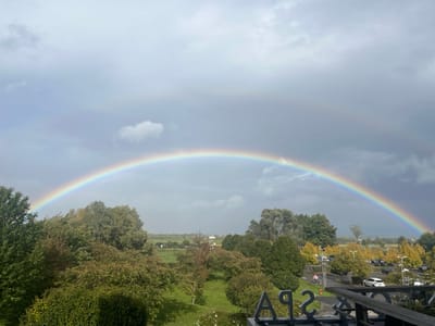 This image captures an elevated outdoor perspective from Ribeauvillé, France, under an overcast sky. A vibrant primary rainbow arches prominently across the upper two-thirds of the frame, extending from the left to the right horizon. A fainter, secondary rainbow is discernible above the primary arc, particularly on the right side. In the immediate foreground, part of a building structure is visible, featuring large, dark gray letters spelling "SPA" in the lower center, and a dark railing on the far right.
The midground is dominated by a diverse landscape of deciduous trees with green and autumnal yellow/brown foliage, bordering a well-maintained grassy area. To the right of the trees, a paved parking lot is visible, containing multiple parked vehicles, including a white SUV/sedan, and several lampposts. At least two indistinct human figures are observed walking on a paved path or near the parking lot in the mid-distance. The background extends into flat, open fields with a few distant structures, indicating a semi-rural environment. No specific interactions between individuals are discernible.