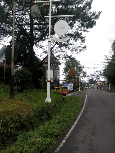 The scene depicts a roadside view in the city of Deothang, Bhutan, under an overcast sky. In the foreground, a prominent white street lamp post with gold accents stands on a grassy incline to the left of an asphalt road. An octagonal lantern is mounted at the top of the post, and a circular white sign bearing Arabic script ("الْعَدُو") and Latin script ("AL ALDU") hangs below it. Behind the post, a multi-story building with a stone-like facade and a brown tiled turreted roof is visible amidst dense green foliage and tall trees. To the right of the street lamp, a dark asphalt road with a solid white edge line extends into the distance. A yellow diamond-shaped road sign and a smaller white sign with '350' in blue are visible on the roadside. Two yellow utility vehicles, one with a pink rear section, are parked on the dirt shoulder next to the road. Further down the road, additional buildings, vehicles, and sparse traffic, including a motorcycle, are discernible. The environment features lush green grass, bushes, and numerous trees, indicating a verdant setting. No specific people or explicit actions are clearly visible.
