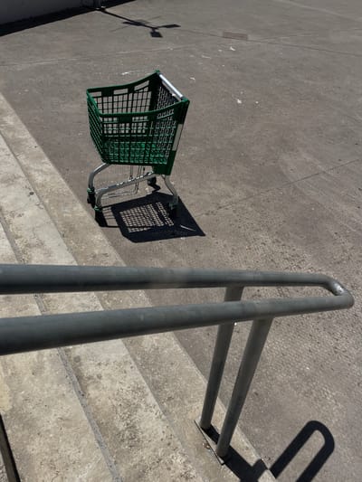 A shopping cart, predominantly green with silver metal framing, rests on a concrete surface. The cart is positioned at an angle, with its basket facing upwards. A dark shadow is cast to the right of the cart, indicating a strong light source, likely the sun. Adjacent to the cart, a metal railing with two parallel bars runs diagonally across the frame. The railing is also casting a shadow. The ground is a rough, textured concrete, with some visible cracks and lighter patches. In the background, further concrete surfaces and what appear to be steps are visible. The scene is outdoors, under bright sunlight, suggesting it is daytime. There are no people visible in the image. The location is Algeciras, Spain.