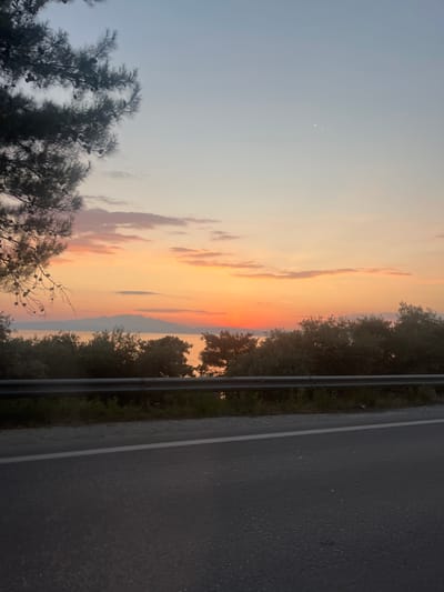 The image shows a sunset over the sea, with trees and a road in the foreground. The sky is filled with shades of orange, pink, and yellow, with a few clouds scattered throughout. The sea is calm and reflects the colors of the sunset. The trees are silhouetted against the sky, and the road is dark and wet, suggesting recent rain. A thin crescent moon is visible in the upper left portion of the sky. The overall mood of the image is peaceful and serene.

The location is Skala Rachoniou, Greece, a coastal town on the island of Thassos. The image appears to have been taken in the evening, as the sun is setting. The weather is clear, with a gentle breeze, as indicated by the movement of the trees. The water is calm, suggesting a tranquil environment.

No people are visible in the image. The objects include trees, a road, a guardrail, and the sea. The scene is a natural landscape with man-made elements. The setting is a roadside overlooking the sea at sunset.

No text is visible in the image.