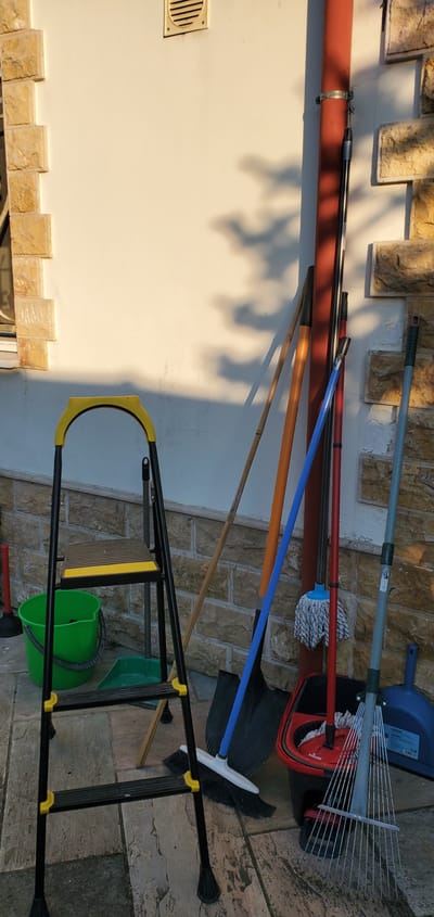 This image shows a collection of cleaning tools leaned against a textured, light-colored wall, partially illuminated by sunlight casting shadows. Among the items are a light blue dustpan, a rake, a mop with a white string head and a red bucket, a blue-handled broom, a shovel, and several long-handled brooms or poles of various colors including red, orange, and natural wood. Some of these tools are propped up by a metal ladder with yellow accents, and a green bucket is visible beneath the ladder. A ventilation grate is mounted on the wall. The scene appears to be outdoors, possibly in a yard or patio area in Pantelimonas, Greece, given the stone-like texture of the ground and wall. The lighting suggests it is daytime, with the sun casting distinct shadows. There is no visible text in the image.