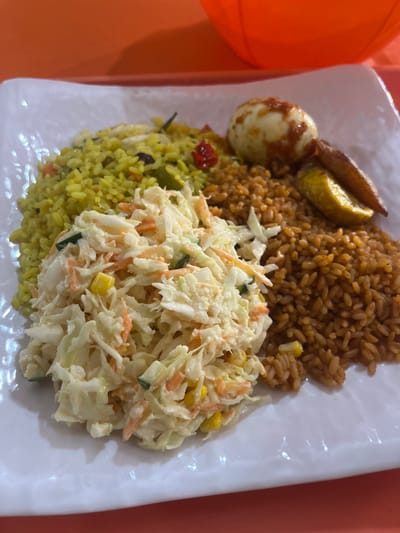 A white rectangular plate, exhibiting a textured surface, rests upon an orange table or tray in Isasi, Nigeria. The plate holds a prepared meal featuring multiple distinct components. A mound of yellow rice, mixed with green and red vegetable fragments, occupies the upper left section. Adjacent to it, on the upper right, is a portion of reddish-brown rice composed of individual grains. A peeled, halved hard-boiled egg topped with a red sauce is positioned above the reddish-brown rice, alongside two slices of fried plantain. The central and lower portion of the plate is filled with coleslaw, consisting of shredded white cabbage, orange carrots, and yellow corn kernels, coated in a creamy, pale dressing. A segment of an orange plastic container is partially visible in the upper right background.