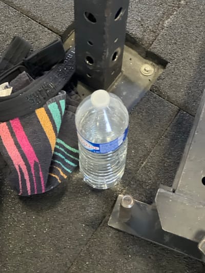 The scene depicts a small section of a fitness facility located in Wolfisheim, France. The floor is covered with dark gray, textured rubber matting. In the foreground, positioned centrally, is a clear plastic bottle of Cristaline natural mineral water, marked 1.5L, appearing mostly full and capped with a white screw lid. To the immediate left of the water bottle, a dark gray towel with horizontal stripes in shades of pink, orange, and teal is folded. Partially obscured behind the towel is a black weightlifting belt, exhibiting a slightly wet or textured surface. To the right and rear of the water bottle, a section of black metal gym equipment is visible. This equipment features a vertical square post with multiple circular adjustment holes, securely anchored to the floor via a bolted base plate. Another small, cylindrical metal peg or fixture is observed on the floor to the lower right. No individuals are present in the frame.