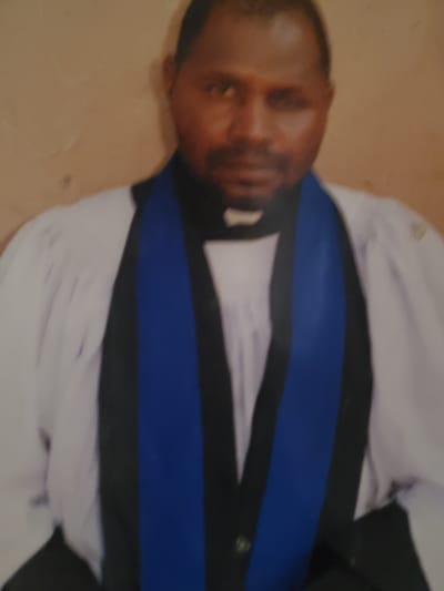 The image is a portrait of a man in a church. He is wearing a white robe and a blue and black stole. The man is looking down and appears to be in thought. The background is a plain wall. The lighting is soft and even. There is no visible text in the image. The man's attire suggests he is a member of the clergy. The overall mood is solemn and reflective.
